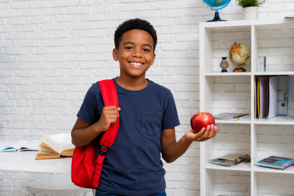 Menino feliz, com mochila nas costas e segurando uma maçã. como preparar seu filho para a volta às aulas de inglês? Foto/Reprodução: Freepik e ChatGPT.